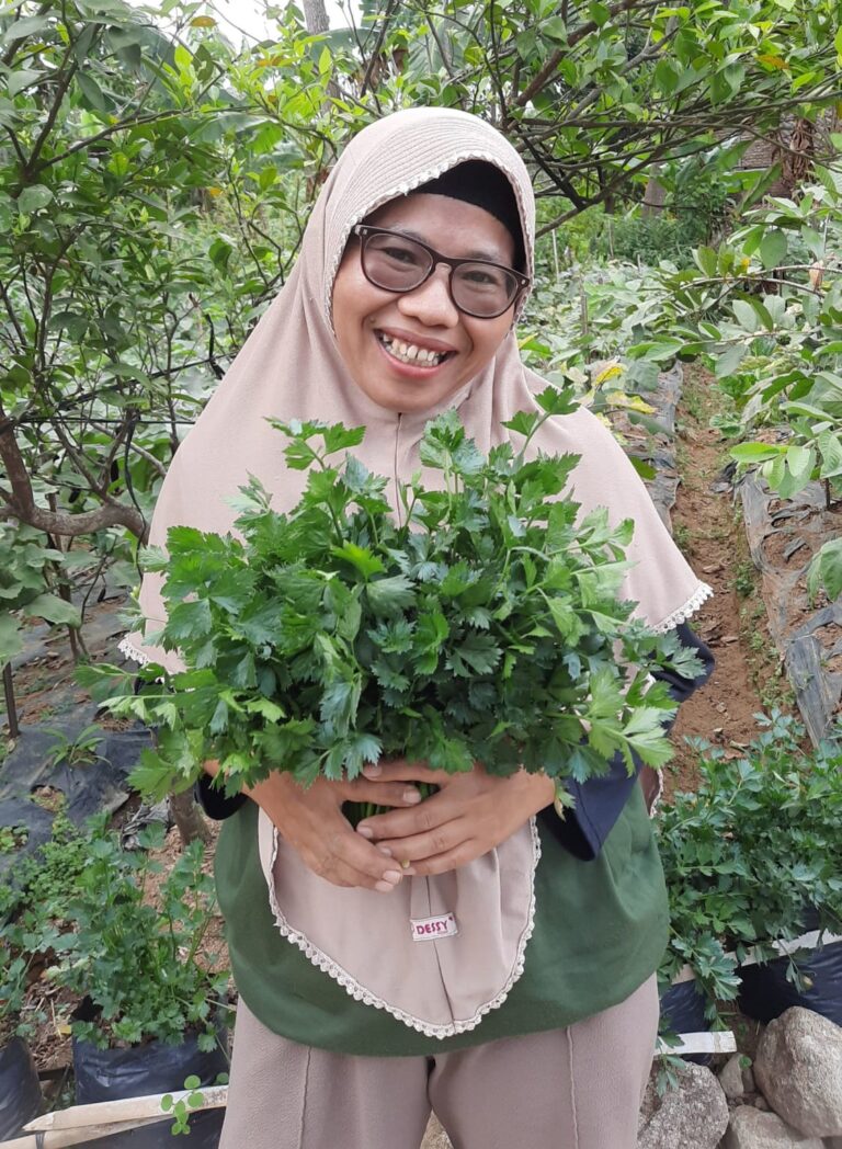 A woman in a headscarf and glasses smiling and holding a bunch of green vegetable and herb leaves