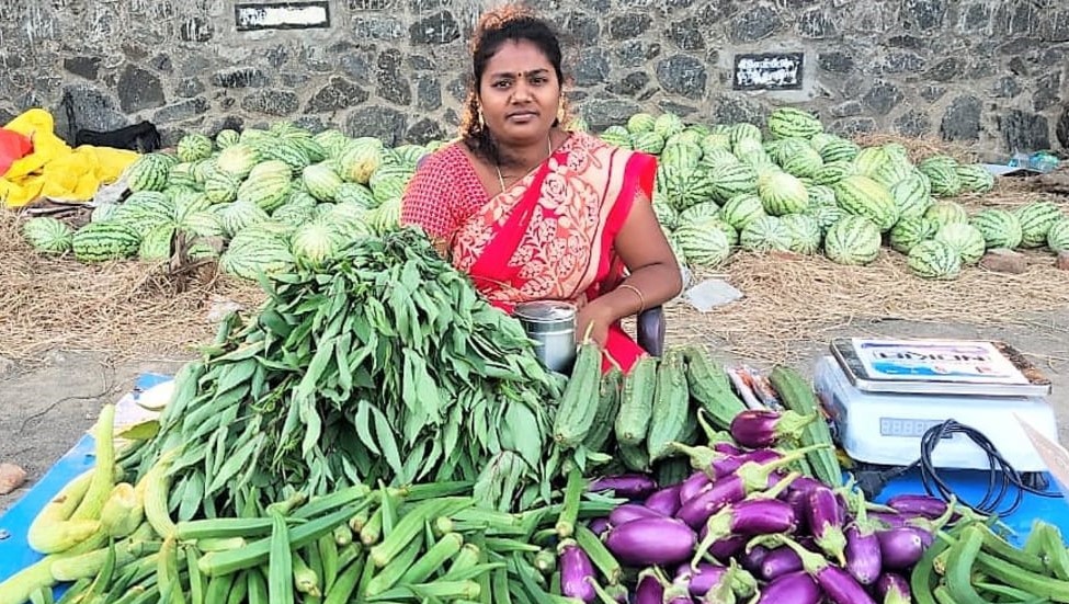 A woman in a sari sitting in front of a large pile of vegetables and a weighing scale.