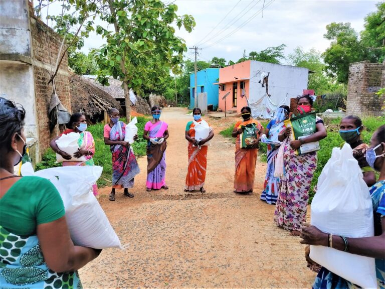 A group of women wearing saris and face masks stand in a circle holding bags of rice.